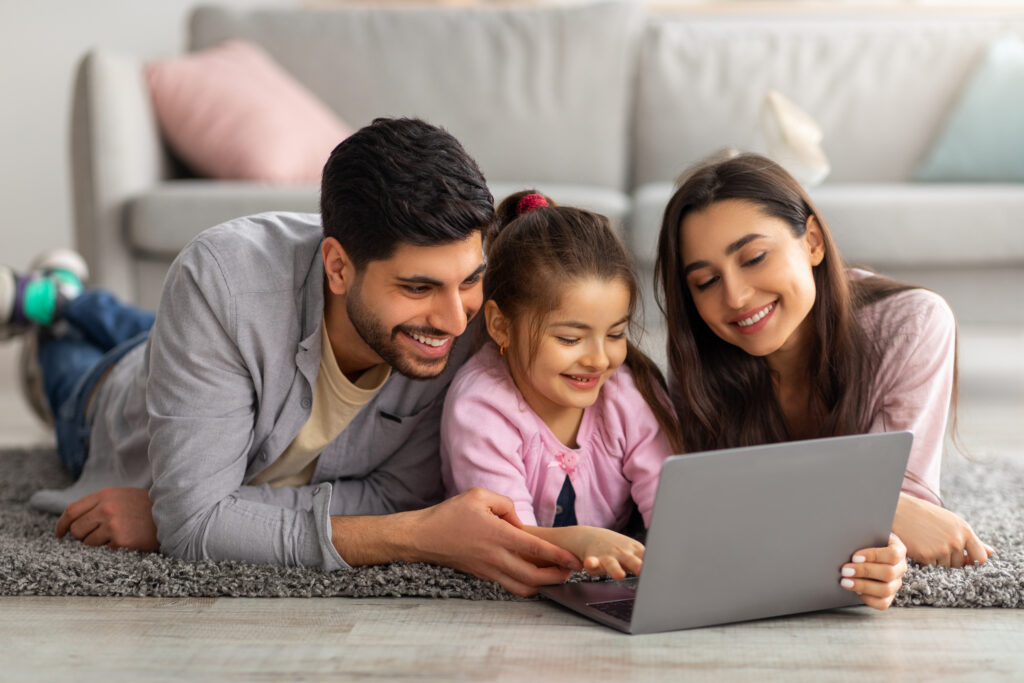 Family weekend. Cheerful arab parents teaching their little daughter using laptop, spending time at home together. Father, mother and girl searching cartoon in internet, lying on the floor carpet